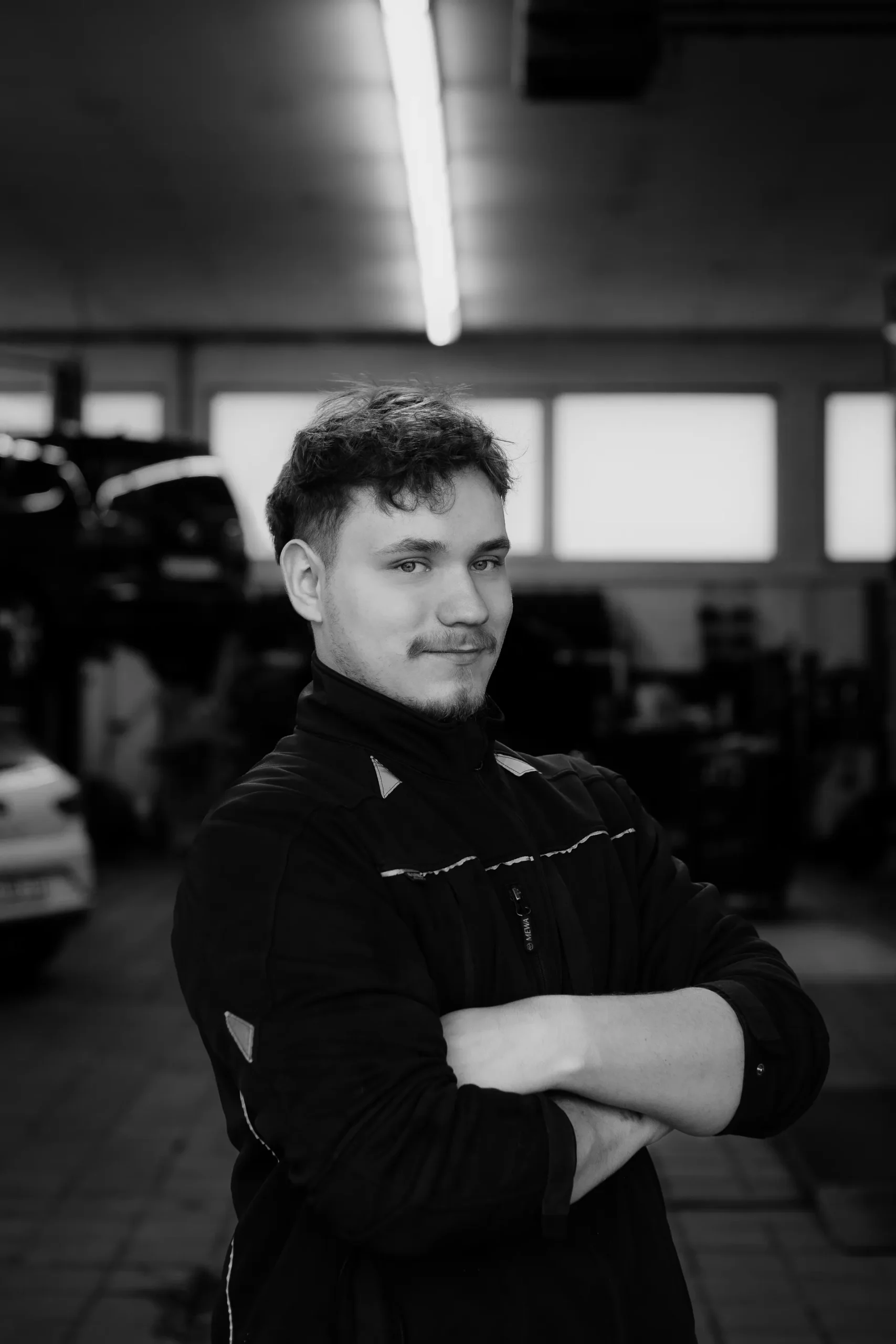 Porträt eines jungen Mitarbeiters mit verschränkten Armen vor Werkstatt-Hintergrund.