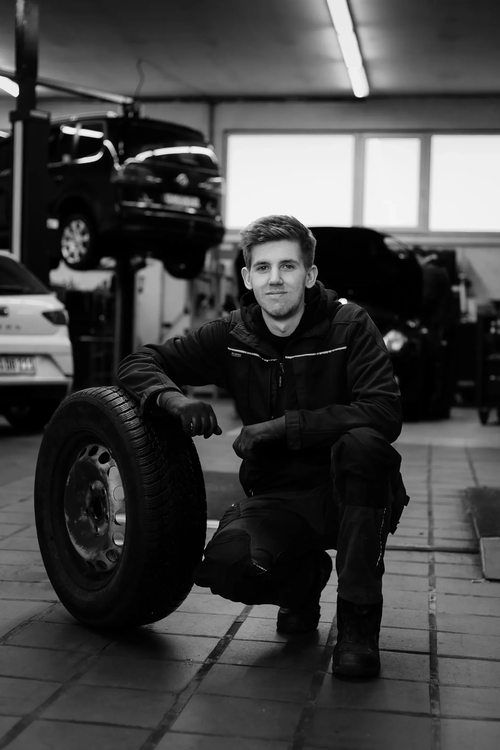 Junger Mitarbeiter kniet lächelnd neben einem Autoreifen in der Werkstatt.